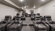 Located at 704 First Dr NW in Austin, Minnesota, this purpose-built theater room offers a fully immersive cinematic experience within a compact, efficiently designed single-floor layout. The room is configured with multiple rows of plush, dark leather power recliner seats arranged in a tiered formation, each equipped with built-in cup holders and wide armrests for maximum comfort during extended viewing sessions. A ceiling-mounted projector delivers crisp imagery onto a large wall-mounted projection screen positioned at the front of the room, ensuring clear sightlines from every seat. The neutral gray walls and low ceiling with recessed lighting create a dark, atmosphere-enhancing environment ideal for an immersive viewing experience. Durable, patterned carpet tiles in a neutral gray tone cover the flooring throughout, providing both practicality and a polished aesthetic. At the rear of the room, dark wood cabinetry flanks a built-in refreshment counter complete with a microwave and mini-refrigerator, allowing guests to enjoy snacks and beverages without leaving the space. A subwoofer is also present, contributing to the room's audio capabilities. Safety features include a visible fire alarm system. A single entry door provides a clean, controlled access point, completing this well-appointed, self-contained theater experience.