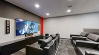 Located at 425 Ballantrae Drive in Winnipeg, this purpose-built cinema room delivers a true home-theatre experience within a compact, efficiently designed single-floor layout. The space is anchored by a large wall-mounted display set against a dark acoustic panel backdrop, flanked by a bold red accent wall that adds a cinematic energy to the room. A surround sound speaker system and a soundbar beneath the screen ensure an immersive audio environment for every viewing.

Seating is arranged in a tiered configuration of plush black leather recliners, providing clear sightlines to the screen from every position. The patterned grey carpet adds texture underfoot while complementing the room's neutral grey walls and dark trim. Recessed ceiling lighting allows for adjustable ambiance, while a wall-mounted ductless HVAC unit ensures year-round climate comfort.

Along the rear and side walls, a built-in wet bar counter with under-counter storage and a dedicated beverage fridge offers convenient refreshment access without leaving the room. A sleek bar-height counter with stools provides an additional seating option. Smart control panels are mounted near the entrance, enabling centralized management of the room's systems. The overall design is modern, polished, and purpose-driven — ideal for private screenings or group entertainment.