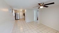 This single-story space features an open-concept layout that seamlessly connects the living area and kitchen, creating a functional and airy feel. Light-colored ceramic tile flooring runs throughout, contributing to a clean, bright aesthetic. The living area is equipped with a ceiling fan with integrated lighting, providing both comfort and illumination.

The kitchen is positioned at the rear of the open space and includes white cabinetry, a stainless steel refrigerator, and a stainless steel range. A track lighting fixture overhead ensures the kitchen is well-lit. A mosaic tile backsplash adds a subtle decorative element behind the cooking area, offering a contrast to the otherwise neutral palette. A window near the kitchen allows natural light to filter into the space.

White walls and white-painted trim are consistent throughout, reinforcing the bright and open atmosphere. A doorway visible from the main living area leads to an additional room, suggesting a practical and connected floor plan. The textured ceiling and simple finishes give the space a straightforward, functional character well-suited for everyday living.