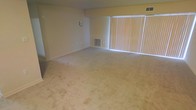 This single-floor space presents a straightforward and functional layout centered around a generously sized living area. The room is finished with wall-to-wall carpet in a neutral beige tone, providing a soft underfoot feel throughout the space. A prominent feature of the living area is the large window fitted with vertical blinds, which allows for adjustable natural light and lends the room an open, airy quality. The light-colored walls complement the neutral flooring, contributing to a bright and cohesive atmosphere. A wall-mounted HVAC vent is positioned near the base of the wall, ensuring climate control throughout the space, while a ceiling vent above adds to the functional infrastructure. A light switch plate and a small wall-mounted fixture are visible, indicating practical utility placements throughout. The overall design is simple and unadorned, offering a blank canvas that adapts easily to a variety of furniture arrangements and décor styles. The space is well-suited for those seeking a clean, no-frills environment with an emphasis on practicality and ease of use.