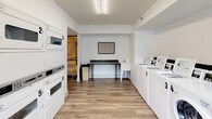 This shared laundry room is designed for practical, efficient use and maintains a clean, organized appearance throughout. The space features white-painted walls and durable wood-look flooring that contribute to a bright, tidy atmosphere. Multiple top-loading washing machines are lined along one wall, each clearly numbered for easy identification, alongside a front-loading washer for added versatility. Stacked dryer units are positioned along the opposite wall, maximizing the use of vertical space and providing ample drying capacity. A dark-topped folding table with seating is centrally located, offering a convenient area for sorting and folding laundry. A wall-mounted notice board keeps users informed of any relevant guidelines or updates. Natural light filters in through horizontal windows along one wall, enhancing the overall brightness of the room. A fire extinguisher is visibly mounted for safety. The layout is thoughtfully arranged to allow comfortable movement between machines and the folding station, making the space functional and easy to navigate. Overall, this laundry room is well-maintained and efficiently organized to serve the needs of its users.