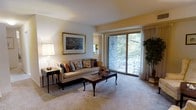 This single-floor model residence offers a thoughtfully designed living space that balances comfort and classic elegance. The living room serves as the heart of the home, featuring wall-to-wall carpeting that adds warmth underfoot and a neutral color palette that creates a calm, inviting atmosphere. A large sliding glass door floods the room with natural light and provides direct access to a private balcony, where mature trees and greenery create a serene outdoor backdrop. The living room is generously proportioned, offering ample space for comfortable seating arrangements, as evidenced by the classic sofa adorned with decorative pillows and a traditional wood coffee table. A framed artwork piece anchors one wall, adding a refined, curated touch to the space. A side table with a classic lamp provides warm ambient lighting, enhancing the room's cozy ambiance. Sheer curtains flank the sliding doors, allowing residents to control light while maintaining the connection to the outdoors. The overall design aesthetic leans toward timeless and traditional, making this model an excellent representation of the comfortable, well-finished living experience available within this community.