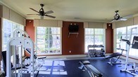 This fitness center is designed to provide a functional and comfortable workout environment. The space is anchored by bold terracotta-red accent walls that add energy and warmth to the room, complemented by white crown molding and trim that lend a polished, finished appearance. Two large windows with fabric valances flank the feature wall, flooding the interior with abundant natural light and offering views of lush outdoor greenery and a fenced exterior area, creating an open and inviting atmosphere. A ceiling fan mounted overhead ensures steady air circulation throughout the workout space. The flooring consists of durable rubber matting, ideal for high-impact exercise and equipment placement. Fitness amenities include a multi-rack dumbbell station stocked with a wide range of free weights, a weight bench, and a strength training machine, providing options for both resistance and functional training. A wall-mounted flat-screen television is positioned centrally on the accent wall, offering entertainment during workouts. The space is thoughtfully arranged to allow for ease of movement between equipment, making it practical for individual use. The combination of natural light, quality equipment, and carefully chosen design details creates a motivating and well-appointed fitness environment.