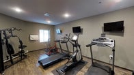 This single-level fitness room provides a practical and well-organized space for cardiovascular exercise. The room features hardwood-style flooring throughout, lending a clean and modern aesthetic to the workout environment. Equipment includes at least two treadmills, an elliptical trainer, and a stationary exercise bike, all arranged to allow comfortable use without overcrowding. Multiple wall-mounted flat-screen televisions are positioned at eye level, enabling users to watch content while exercising. A window along one wall allows natural light to filter into the space, contributing to a more open and energizing atmosphere. The neutral-toned walls and standard ceiling height keep the environment straightforward and functional. Protective floor mats are placed beneath the cardio machines to preserve the flooring and reduce equipment vibration. A towel rack or display board is also visible near the window wall, adding a practical amenity. Overall, the space is efficiently laid out, prioritizing usability and accessibility for fitness-focused occupants.