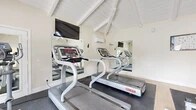 Located at 1200 Fairhaven Ave in Santa Ana, California, this fitness center is a well-appointed gym space designed to support a comprehensive workout routine. The room is immediately distinguished by its soaring vaulted ceiling with exposed white-painted wood beams and a matching shiplap ceiling deck, lending the space an airy, open feel despite its compact footprint.

The gym is equipped with multiple Star Trac treadmills, an elliptical trainer, a recumbent stationary bike, and a multi-station strength machine, offering a balanced mix of cardiovascular and resistance training options. Rubber flooring with a speckled finish covers the workout areas, providing both safety and durability underfoot, while a section of wood-look flooring adds a warm visual contrast.

Large mirrors are strategically placed along the walls to enhance the sense of space and allow users to monitor their form. A wall-mounted flat-screen television provides entertainment during cardio sessions, and a wall-mounted paper towel dispenser supports cleanliness. A mini-split air conditioning unit ensures climate comfort year-round.

Natural light fills the space through a set of full-length glass French doors that open directly to an outdoor pool area, creating a seamless connection to the exterior and providing refreshing views of the surrounding landscaped grounds.