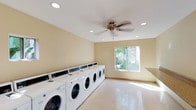 The Laundry Center at 6675 Jaffe Ct in San Diego is a well-designed, single-floor communal facility built for convenience and functionality. The space is finished with warm, neutral-toned walls and a clean, polished composite floor that reflects natural light throughout the room.

The main washing area lines one wall with a full row of front-load washing machines, offering ample capacity for multiple users simultaneously. Opposite the washers, a bank of stacked commercial dryers provides efficient drying options, maximizing the use of floor space. A granite-topped folding counter runs along one side of the room, offering a generous surface for sorting and folding laundry.

Natural light enters through multiple windows, including a large picture window and a row of smaller clerestory windows, giving the space an open and airy feel. A ceiling fan with recessed lighting overhead ensures consistent air circulation and comfort year-round. The glass entry door opens to an outdoor walkway bordered by greenery, contributing to the pleasant atmosphere.

Practical amenities include a vending or detergent dispenser mounted on the wall, as well as clearly placed recycling and waste bins. The layout is thoughtfully organized to allow comfortable movement throughout the space, making this laundry center a clean, efficient, and user-friendly facility for the surrounding community.