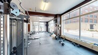 Located at 421 19th St in Moline, Illinois, this fitness facility spans a single floor and offers a well-rounded workout environment divided into distinct functional areas. The space is characterized by exposed dark wood beams that contrast with light gray walls and white ceilings, lending a clean, modern-industrial aesthetic throughout.

The primary workout area is a spacious, open floor plan covered in durable interlocking rubber tiles. It is comprehensively equipped with free weights including barbells, dumbbells, and weight plates, as well as a cable machine, benches, a rowing machine, and stability balls. Large floor-to-ceiling windows along one exterior wall flood the space with abundant natural light and offer direct views of the surrounding urban streetscape.

A dedicated cardio room features carpeted flooring and houses a treadmill, elliptical trainer, stationary bikes, and a wall-mounted television—all positioned to benefit from the natural light streaming through a large exterior window.

A welcoming reception and lounge area connects the two workout zones, furnished with a tufted bench-style sofa, a storage credenza, and a water cooler. Decorative artwork on the walls adds a personalized touch. The facility also includes a private restroom accessible via a rich wood-trimmed door. Overhead fluorescent lighting ensures consistent illumination throughout all areas of the space.