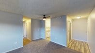 This single-floor space features an open-concept layout that connects the main living area to the kitchen and adjoining rooms. The living area is finished with wall-to-wall carpeting in a neutral tone, contributing to a comfortable and understated atmosphere. Light grey walls run throughout, providing a clean, cohesive aesthetic that pairs well with the white trim and baseboards.

A ceiling fan with an integrated light fixture is centrally mounted above the living space, offering both ventilation and ambient lighting. The textured popcorn ceiling adds a classic residential character to the space.

The kitchen is visible through an open passageway and features white cabinetry, a sink, and light wood-style flooring, creating a functional and bright cooking area. Adjacent to the kitchen, a doorway leads to an additional area with matching wood-style flooring, likely a utility or entryway space.

A hallway extends from the living area toward what appears to be additional rooms, with doors visible along the corridor. The layout is practical and efficiently organized, offering clear sightlines between the main living zone and supporting spaces throughout the unit.