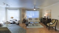 This single-floor residence features an open-concept layout that seamlessly connects the living and dining areas, creating a comfortable and functional living space. Warm hardwood flooring extends throughout, adding a cohesive and inviting feel to the interior.

The living area is anchored by a light-toned sofa accented with decorative pillows, complemented by a glass-top coffee table and a small upholstered ottoman. A dark wood dresser with multiple drawers lines one wall, providing ample storage. A ceiling fan with an integrated light fixture is centrally positioned, promoting air circulation and adding a practical touch to the space.

Natural light is a defining feature, with multiple windows dressed in white curtains and horizontal blinds allowing generous brightness throughout the day. The dining area, positioned to one side, accommodates a small round table with seating for two, ideal for casual meals.

Neutral wall tones in soft gray and beige contribute to the calm, understated ambiance of the space. Overall, the layout is efficiently designed, making thoughtful use of the available space while maintaining an open and airy atmosphere.