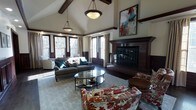 This single-floor residence showcases exceptional craftsmanship and thoughtful design throughout. The centerpiece of the home is a grand living area distinguished by soaring vaulted ceilings adorned with rich, dark-stained exposed wood beams that create a striking architectural contrast against the light, neutral walls. Elegant pendant lighting hangs from the peak of the vault, casting a warm glow across the space and complementing the dark hardwood flooring below.

A substantial wood-surround fireplace anchors one wall of the living area, featuring classic craftsman-style detailing and dark millwork consistent with the home's design language. A large piece of abstract artwork is displayed prominently above the mantel, adding a contemporary accent to the traditional framework.

An array of windows with dark wood trim lines the perimeter of the main living space, flooding the room with natural light and offering views of the surrounding mature trees. Wainscoting and crown molding details further reinforce the home's refined character.

The living area is generously proportioned, comfortably accommodating multiple seating arrangements including a plush sectional, a floral accent chair, and a glass-topped oval coffee table centered on a large area rug. The overall ambiance is warm, inviting, and timelessly elegant.