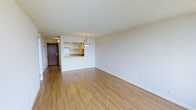 This single-floor space offers an open and airy living area finished with warm-toned hardwood-style flooring that extends throughout the main living zone, creating a cohesive and inviting atmosphere. The walls are painted in crisp, neutral white tones, lending a clean and versatile backdrop suitable for a variety of interior styles.

A notable architectural feature is the pass-through kitchen opening, which connects the kitchen to the main living and dining area. This design element promotes an open-concept feel while maintaining a defined kitchen space. Light wood cabinetry is visible through the pass-through, complementing the flooring tones and contributing to the overall warmth of the space. A pendant light fixture hangs above the pass-through counter, providing both functional task lighting and a subtle decorative touch.

The textured ceiling adds depth overhead, while white baseboards provide a clean transition between the walls and flooring. A solid wood entry door is visible toward the rear, offering a classic contrast to the lighter finishes throughout. The overall ambiance is bright, neutral, and well-suited for comfortable everyday living.