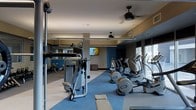 This fitness center is a thoughtfully designed, single-floor facility built to support a comprehensive workout experience. The space is outfitted with a robust selection of cardio equipment, including a row of elliptical trainers positioned along the perimeter to take advantage of the large windows that flood the room with natural light and provide views of the surrounding exterior. Strength training machines are prominently featured, offering users a variety of resistance-based exercise options. A dedicated free weights and stretching area is visible toward the rear, complemented by full-length mirrors that enhance the sense of space and allow for proper form checks during workouts. The interior design features a modern color palette, with deep blue accent walls contrasting against light-toned flooring and neutral ceiling finishes. Ceiling fans are mounted overhead, contributing to air circulation throughout the space. A wall-mounted flat-screen television provides entertainment for users during their sessions. Recessed and ambient lighting, combined with the generous natural light from the windows, creates a bright and energizing atmosphere well-suited for fitness activity.
