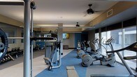 This fitness center is a thoughtfully designed, single-floor facility built to support a comprehensive workout experience. The space is outfitted with a robust selection of cardio equipment, including a row of elliptical trainers positioned along the perimeter to take advantage of the large windows that flood the room with natural light and provide views of the surrounding exterior. Strength training machines are prominently featured, offering users a variety of resistance-based exercise options. A dedicated free weights and stretching area is visible toward the rear, complemented by full-length mirrors that enhance the sense of space and allow for proper form checks during workouts. The interior design features a modern color palette, with deep blue accent walls contrasting against light-toned flooring and neutral ceiling finishes. Ceiling fans are mounted overhead, contributing to air circulation throughout the space. A wall-mounted flat-screen television provides entertainment for users during their sessions. Recessed and ambient lighting, combined with the generous natural light from the windows, creates a bright and energizing atmosphere well-suited for fitness activity.