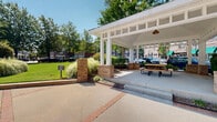 Located at the center of a walkable mixed-use district in Holly Springs, North Carolina, this outdoor public space offers a well-designed gathering area that blends green landscaping with thoughtful hardscape elements. A white-framed covered pavilion anchors one side of the space, featuring open-air seating with picnic tables beneath pendant lighting — ideal for community gatherings or casual breaks throughout the day. Brick-accented concrete pathways crisscross the area, creating a structured yet inviting layout that connects the pavilion to surrounding sidewalks and green spaces. The central lawn is expansive and well-maintained, bordered by mature shade trees, ornamental grasses, and mulched planting beds that lend a polished, park-like character to the surroundings. Metal benches are positioned along the perimeter, offering shaded seating beneath the tree canopy. Brick pillar accents and directional signage mark entry points throughout, adding to the cohesive streetscape design. The space is framed by multi-story brick commercial and mixed-use buildings featuring green awnings and ground-floor retail, creating a lively urban backdrop. Street-level parking and wide sidewalks make the area highly accessible, reflecting a pedestrian-friendly design philosophy central to the Holly Springs Town Center.