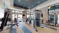 This modern fitness center is thoughtfully designed to support a comprehensive workout experience. The space features a striking tray ceiling finished in a deep teal-blue tone, accented by recessed lighting and stylish pendant lights that create a polished, welcoming ambiance. Large windows positioned along the walls flood the facility with natural light, while wall-mounted flat-screen TVs provide entertainment throughout the workout.

The layout is divided into distinct functional zones. The cardio area is equipped with multiple treadmills and elliptical machines arranged in a row, all facing a bank of windows. Adjacent to this is a dedicated strength training section featuring a multi-functional cable machine, weight benches, and a squat rack. A well-stocked dumbbell rack offers a full range of free weights for varied resistance training.

Full-height framed mirror panels run along one wall, visually expanding the space and allowing users to monitor their form. The flooring combines large-format tile with rubber sport flooring in the equipment zones, providing both durability and safety. A hydration station with dual water fountains and cabinetry offers convenience, while a glass-paneled entryway provides a bright, open transition into the space. The overall design balances functionality with a contemporary aesthetic.