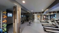 This well-appointed fitness center offers a thoughtfully designed workout space that balances functionality with contemporary style. The interior is characterized by striking wood-panel accent walls in warm, rustic tones that contrast beautifully against the cool gray walls and ceiling, creating a modern yet inviting atmosphere.

The cardio zone is generously equipped with a variety of machines, including treadmills, stationary cycling bikes, and an elliptical trainer, catering to a range of fitness preferences and workout routines. Full-length mirrored walls line the space, enhancing the sense of openness while allowing users to monitor their form during exercise.

Recessed ceiling lighting provides bright, even illumination throughout the floor, ensuring a well-lit environment at all times. The flooring features durable gray rubber or sport-grade material, practical for both equipment use and high-traffic movement. A wall-mounted bulletin board is visible near the entrance, likely used for facility information or community notices.

The single-floor layout is efficiently organized, making navigation intuitive and the overall experience streamlined. The combination of modern finishes, ample equipment, and thoughtful design details makes this fitness center a polished and practical facility for daily use.
