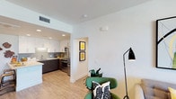 This model unit at 986 Broadway in Chula Vista showcases a thoughtfully designed, single-floor open-concept layout that seamlessly blends functionality with contemporary style. The living area features clean white walls, light wood-style flooring, and an abundance of natural light, creating a warm and welcoming ambiance. A modern floor lamp and carefully curated accent pieces, including a green lounge chair with a patterned throw pillow, add a touch of personality to the space. Framed yellow artwork adorns the hallway wall, contributing a vibrant pop of color to the neutral palette. The kitchen is a standout feature, boasting white upper cabinetry contrasted by rich dark lower cabinets, stainless steel appliances including a refrigerator and range, recessed lighting, and a clean subway tile backsplash. A peninsula provides additional prep space and casual seating. The open sightlines between the kitchen and living area enhance the sense of spaciousness throughout the unit. Recessed and ceiling-mounted lighting fixtures ensure every corner is well-lit, while the crisp white ceilings contribute to an airy, modern feel throughout this well-appointed Chula Vista residence.