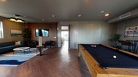 This well-appointed entertainment lounge is designed with a modern, urban aesthetic that blends functionality with sophisticated style. Throughout the space, herringbone-patterned hardwood flooring provides a warm, cohesive foundation, while richly textured cork wall panels and subtly striated gray wall coverings create a layered, tactile atmosphere. Recessed ceiling lighting illuminates each zone evenly, contributing to the polished overall ambiance.

The main lounge area features a pool table with a solid wood frame and black felt surface, complemented by a seating nook with a wall-mounted flat-screen television, black leather armchairs, and a Jenga tower for casual entertainment. A separate lounge section offers plush sofas, high-backed accent chairs, and curated artwork, creating an inviting social setting. An additional game room includes an air hockey table, a sofa, and generous natural light from multiple windows.

A dedicated bar and kitchenette area is thoughtfully equipped with dark stone countertops, matte gray cabinetry, a stainless steel sink, a wine refrigerator, and built-in appliances, alongside open shelving ideal for entertaining. A bistro-style nook with high-top tables and leather bar chairs frames a large picture window showcasing panoramic city skyline and mountain views, making it a standout feature of the space.
