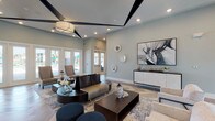 This single-story space presents a polished, contemporary design aesthetic with a cohesive coastal-inspired palette. The open main lounge area is anchored by striking herringbone-patterned flooring in a light wood tone, which extends throughout the space and contributes to a sense of expansive flow. The elevated ceilings feature bold geometric black beam accents, adding architectural interest and visual depth overhead, while recessed lighting and statement pendant fixtures provide layered illumination.

The lounge is furnished with a curated mix of modern seating, including sculptural dark-framed sofas, upholstered accent chairs, navy blue ottomans, and dark wood coffee tables styled with decorative objects. A media console along one wall is paired with large-scale artwork and circular decorative mirrors, adding dimension to the soft blue-grey walls.

Multiple sets of glass-paned French doors line the perimeter, flooding the interior with natural light and offering direct views of an outdoor pool area with tropical landscaping. A glass-walled office or leasing area is visible near the entrance, furnished with workstations and a chandelier. Additional rooms accessible via interior doors appear to serve as private meeting or activity spaces. The overall ambiance is refined, light-filled, and welcoming.