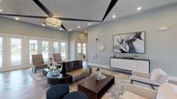 This single-story space presents a polished, contemporary design aesthetic with a cohesive coastal-inspired palette. The open main lounge area is anchored by striking herringbone-patterned flooring in a light wood tone, which extends throughout the space and contributes to a sense of expansive flow. The elevated ceilings feature bold geometric black beam accents, adding architectural interest and visual depth overhead, while recessed lighting and statement pendant fixtures provide layered illumination.

The lounge is furnished with a curated mix of modern seating, including sculptural dark-framed sofas, upholstered accent chairs, navy blue ottomans, and dark wood coffee tables styled with decorative objects. A media console along one wall is paired with large-scale artwork and circular decorative mirrors, adding dimension to the soft blue-grey walls.

Multiple sets of glass-paned French doors line the perimeter, flooding the interior with natural light and offering direct views of an outdoor pool area with tropical landscaping. A glass-walled office or leasing area is visible near the entrance, furnished with workstations and a chandelier. Additional rooms accessible via interior doors appear to serve as private meeting or activity spaces. The overall ambiance is refined, light-filled, and welcoming.