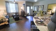 This single-floor residence showcases a thoughtfully designed open-concept layout that connects the kitchen, dining, and living areas into one cohesive, flowing space. Rich, dark hardwood flooring runs throughout, anchoring the interior with warmth and sophistication. The kitchen features a curved granite-topped island with bar seating, providing both a functional prep area and a casual dining option. Adjacent to the kitchen, a dedicated dining area accommodates a round table with seating, framed by vibrant artwork that adds a pop of color to the neutral wall tones. The living area is generously proportioned, furnished with low-profile dark wood accent tables atop layered area rugs, creating an inviting lounge atmosphere. A cozy accent chair occupies a sunlit corner near a tall window dressed with floor-length curtains, while a decorative dresser and table lamp add warmth to the space. Multiple windows fitted with blinds allow natural light to filter throughout, enhancing the open and airy ambiance. Carefully placed greenery, decorative vases, and curated artwork contribute to an overall aesthetic that is both polished and welcoming.