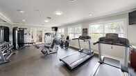 Located at 202 Saint James Ave in Goose Creek, South Carolina, this fitness center offers a well-equipped exercise space designed to support a comprehensive workout routine. The room features durable rubber-composite flooring throughout, providing both safety and functionality underfoot. Crown molding and neutral gray walls lend a clean, polished aesthetic to the space.

Natural light pours in through multiple large windows along the exterior walls, offering views of the surrounding neighborhood and creating an open, inviting atmosphere. Recessed lighting and flush-mount ceiling fixtures supplement illumination throughout the room.

The facility is outfitted with a diverse selection of Life Fitness cardio equipment, including treadmills, an elliptical trainer, and a recumbent exercise bike. Strength training is well represented with a multi-function cable machine, a leg press/extension unit, and a dedicated dumbbell rack stocked with a range of free weights. A full-length wall mirror on one side enhances the sense of space and allows users to monitor their form during workouts. A wall-mounted flat-screen television provides entertainment during exercise sessions.

French doors connect the fitness center to an adjacent common area, and a standard exit door provides additional access. The space is efficiently organized to maximize usability while maintaining comfortable circulation between equipment stations, making it a practical and fully functional amenity.
