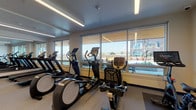 This fitness center is designed to support a comprehensive workout experience within a clean, modern environment. The space is divided into two connected zones, each purpose-built for different types of exercise. The cardio area lines the windowed wall, where a row of treadmills, elliptical trainers, and a stationary bike are positioned to take full advantage of expansive windows that frame outdoor views and flood the room with natural light. Recessed linear lighting runs along the ceiling, ensuring consistent illumination throughout. The flooring throughout is a durable speckled rubber surface, providing both comfort and traction. The secondary zone serves as a functional training and free weights area, featuring a full-length wall mirror that visually expands the space. A tiered dumbbell rack, medicine balls, resistance bands, and a TRX suspension system offer a wide range of strength and conditioning options. White walls with blue accent baseboards give the space a crisp, professional aesthetic. A wall-mounted split air conditioning unit ensures climate control, while a glass-paneled door connects the two areas and maintains an open, accessible flow. The overall ambiance is bright, clean, and motivating.