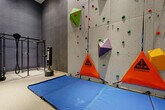 Located at 728 Broadway in Saugus, MA, this purpose-built climbing room is a compact yet fully equipped fitness space designed for indoor bouldering and functional training. The centerpiece is a tall, panel-style bouldering wall that spans the full length of one side of the room, fitted with an array of colorful climbing holds in various shapes and sizes to accommodate different skill levels and route configurations. Safety is well addressed with large blue foam crash mats positioned along the base of the wall, and Strublue "Clip In" safety markers are prominently displayed. The room features impressively high ceilings that allow for a substantial climbing surface, contributing to a challenging and authentic bouldering experience. Complementing the climbing wall, a freestanding functional training rig with cable attachments and a standing punching bag provides additional cross-training options. The flooring consists of a durable, light-colored rubberized surface, and the walls are finished in a sleek dark gray, lending the space a modern, athletic aesthetic. A wooden open-shelf storage unit offers space for gear and accessories. One side of the room features floor-to-ceiling glass panel partitions, offering visibility into adjacent areas while maintaining a clean, professional look. Recessed lighting ensures the space is well-lit throughout.