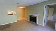 This single-floor residence offers a functional and comfortable layout highlighted by warm, neutral design choices throughout. The main living area is carpeted in a light beige tone and centers around a tile-surround fireplace framed by a classic white wood mantel, with a matching tiled hearth extending onto the floor — a practical and visually grounding focal point. The walls are painted in a soft sage green, contributing to a calm and understated ambiance.

The textured popcorn ceiling adds a traditional touch, and flush-mount ceiling lighting provides general illumination throughout the space. A pass-through opening connects the living area to the kitchen, promoting an open, communicative flow between the two spaces. The kitchen area, visible through the pass-through, features light cabinetry and countertops, suggesting a clean and functional workspace.

Carpeting extends throughout the living and adjoining areas, adding warmth underfoot. HVAC venting is visible in the ceiling, indicating a central climate control system. The overall layout is straightforward and practical, suited to everyday living with a welcoming, low-maintenance aesthetic.