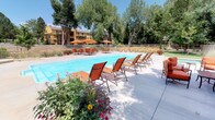 This expansive outdoor amenity space is designed to provide a comprehensive resort-style experience for residents. At its center is a generously sized rectangular swimming pool with crystal-clear blue water, complemented by an adjacent spa, offering options for both active swimming and relaxed soaking. The surrounding pool deck is well-furnished with rows of orange sling-back chaise lounges ideal for sunbathing, while multiple dining tables paired with vibrant orange market umbrellas provide shaded areas for outdoor meals and gatherings. A dedicated lounge zone features cushioned seating arranged around a built-in fire pit, creating an inviting spot for evening relaxation. An outdoor kitchen station, constructed with a stacked stone surround and equipped with a stainless steel gas grill, adds convenient al fresco cooking capability to the space. The entire area is enveloped by mature, full-canopy trees that offer natural shade and a lush, park-like backdrop. A clubhouse building with large windows borders the pool deck, providing direct visual and physical access between indoor and outdoor spaces. The perimeter is secured by a wrought iron fence with gated entry, and colorful flowering plants and manicured green lawn accents complete the well-maintained setting.
