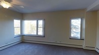 This single-floor space presents a clean, straightforward layout centered around a generously sized main room. The room is finished with neutral, cream-toned walls that create a bright and versatile backdrop, complemented by wall-to-wall carpeting in a muted gray-blue shade that adds a sense of warmth underfoot. Three windows are positioned along the main wall, allowing abundant natural light to fill the interior while offering views of surrounding greenery and trees, enhancing the overall sense of openness. The windows are fitted with roller shades, providing flexible light control. A baseboard heating system runs along the base of the exterior wall, ensuring consistent climate control throughout the space. The ceiling is smooth and painted white, contributing to a clean, uncluttered aesthetic. The room's proportions provide ample open floor space, making it well-suited for a variety of furniture arrangements or functional uses. Overall, the space offers a practical and well-lit environment with a neutral palette that serves as an adaptable foundation for personalization.