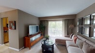 This single-floor residence offers a thoughtfully arranged layout with distinct living spaces finished in warm, neutral tones. The living room serves as a central gathering area, featuring plush carpeting, taupe walls, and a large sliding glass door that opens to a private balcony, filling the space with natural light and offering views of surrounding greenery. A sectional sofa, glass-top coffee table, and wood media console with a flat-screen television complete the room. Decorative wall mirrors add depth to the space.

The dining room is anchored by a glass-top table with seating for six, illuminated by a classic chandelier. A wood-and-glass display cabinet provides an elegant storage feature. Adjacent to the dining area, the kitchen offers granite countertops, honey-toned cabinetry, a gas range, dishwasher, and tile flooring.

The primary bedroom is generously sized with large sliding doors that frame outdoor views, complemented by soft carpeting and ample natural light. A secondary bedroom functions as a dedicated home office, featuring built-in shelving and a large dark-wood desk unit.

The bathroom includes granite countertops, warm wood cabinetry, tile flooring, and a well-lit vanity mirror.