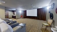 This single-floor screening room is thoughtfully designed to deliver a true cinematic experience. At the front of the room, a large projection screen is framed by floor-to-ceiling rich brown curtains, creating a dramatic focal point that sets the stage for movie viewing. The seating area is furnished with rows of generously sized gray and white upholstered chairs, each equipped with individual side tables, offering both comfort and convenience for guests. The patterned carpet adds warmth and a classic theater aesthetic throughout the space. The walls are painted in a soft gray tone and adorned with decorative film reel wall art in blue, red, and charcoal, reinforcing the cinematic theme. Recessed ceiling lights and wall-mounted sconces provide a warm, ambient glow, complementing the room's intimate atmosphere. A dedicated concession area is tucked to one side, featuring a classic red popcorn machine, dark cabinetry, and a countertop, adding a nostalgic and functional touch. A private entrance door with a wood frame and louvered window provides easy access. The overall ambiance is polished and inviting, blending the comfort of a lounge with the functionality of a professional screening room.