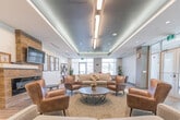 Located at 212 Davis Dr in Newmarket, Ontario, this well-appointed amenity lounge offers a generously sized, single-floor communal space designed for comfort and social interaction. The open-concept layout is anchored by a sophisticated seating area furnished with plush sofas, rich leather armchairs, and a round coffee table, all centered around a sleek linear fireplace set into a rustic wood-clad accent wall. A wall-mounted television above the fireplace adds a modern entertainment focal point, complemented by framed photographic artwork that lends a refined, gallery-like touch to the space. The ceiling features a distinctive tray design with integrated linear and recessed lighting, creating a warm, layered ambiance throughout. Large glazed entry doors allow abundant natural light to pour in from the building entrance, enhancing the open and airy feel of the lounge. Light wood-look flooring runs seamlessly through the space, tying together the contemporary aesthetic. Adjacent to the lounge is a fully equipped kitchen with crisp white cabinetry, a tiled backsplash, stainless steel appliances including a refrigerator, stove, and dishwasher, and a kitchen island with seating — ideal for catering or communal dining. A separate dining nook with upholstered chairs rounds out the functional versatility of this well-designed amenity space.