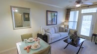 This single-floor residence combines a living area and dining space in an open, functional layout. The living room centers around a light gray upholstered sofa accented with decorative throw pillows, positioned beneath a framed art piece that adds a pop of color to the soft, neutral-toned walls. A dark wood side table flanks the sofa, displaying elegant white ceramic vases that provide a clean, modern accent. Plush carpeting underfoot adds warmth and comfort throughout the space.

Natural light filters in through a window dressed with patterned drapes, complemented by a ceiling fan overhead that enhances airflow and comfort. A dark wood coffee table with a lower shelf anchors the seating area, while a wall-mounted mirror near the dining space adds depth and brightness.

The dining area features a glass-top table surrounded by green upholstered chairs, set with decorative dishware and folded cloth napkins, lending an elegant, curated feel. Together, the spaces share a cohesive, warm palette and thoughtfully chosen furnishings that make the layout feel welcoming and well-appointed.