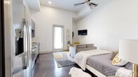 This single-floor studio is designed with a clean, contemporary aesthetic that maximizes both function and style. The open-concept layout seamlessly integrates the sleeping, living, and kitchen areas beneath impressively tall ceilings, creating an airy, expansive feel. A ceiling fan overhead and large windows fitted with blinds allow for adjustable natural light throughout the main living space.

The kitchen is well-appointed with gray shaker-style cabinetry, granite countertops, a white subway tile backsplash, and a full suite of stainless steel appliances, including a refrigerator, range, built-in microwave, and dishwasher. The polished concrete floors extend throughout, adding an industrial yet refined character to the space.

The living and sleeping area is versatile and open, offering ample room for a full bed and sofa arrangement. A wall-mounted television and framed artwork contribute to the modern, curated ambiance. Adjacent to the main area, a dedicated walk-in closet provides built-in shelving and additional storage. The full bathroom features gray cabinetry with granite countertops, a tub and shower combination, and consistent concrete flooring, maintaining the cohesive design language found throughout the space.