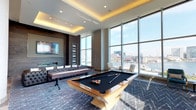 This elegant amenity lounge is defined by its soaring floor-to-ceiling glass walls that frame breathtaking panoramic views of a bustling harbor, marina, and city skyline beyond. Abundant natural light floods the interior throughout the day, creating a bright and inviting atmosphere that seamlessly connects the indoors with the waterfront scenery outside.

The space is thoughtfully divided into distinct recreational and social zones. A central game area features a premium billiards table with a sleek wood frame and a shuffleboard table, both anchored by a boldly patterned chevron area rug. Surrounding this zone, richly upholstered tufted leather sofas and lounge chairs provide comfortable seating, complemented by a wall-mounted flat-screen television and an integrated fireplace set within a dark, textured feature wall.

A separate lounge seating area offers an intimate retreat with stylish gold-accented chairs and chaise lounges positioned directly against the expansive glass facade, ideal for taking in the harbor views. An outdoor terrace is accessible through large glass doors, extending the entertaining space further.

The entryway is grand and airy, with soaring double-height ceilings, large-format tile flooring, and glass vestibule doors that invite natural light deep into the space. Architectural columns and coffered ceiling details with warm ambient lighting add a refined, contemporary character throughout this impressive amenity floor.