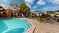 Located at 4083 Sunbeam Rd in Jacksonville, Florida, this outdoor amenity space offers a well-appointed resort-style experience designed for relaxation and recreation. The centerpiece is a generously sized swimming pool with crystal-clear turquoise water, framed by an expansive paver deck that provides ample space for lounge seating and sunbathing. Surrounding the pool, lush tropical landscaping — including vibrant red ti plants, ornamental grasses, and mature shade trees — creates a lush, private atmosphere that evokes a Florida resort setting.

Adjacent to the pool, a separate hot tub area offers a more intimate retreat, complemented by additional paver hardscaping and decorative stone-clad planters. Outdoor grilling stations are conveniently positioned nearby, making the space well-suited for social gatherings. A shaded dining area with umbrella-covered tables and chairs provides a comfortable spot to enjoy the outdoors.

Access to the amenity area is provided through a set of double glass-panel doors with a classic trim surround, offering clear sightlines to the pool from the building interior. The multi-building residential backdrop, featuring warm earth-tone stucco exteriors and balconies, frames the space and contributes to a cohesive community aesthetic. A scenic pond with a fountain is visible beyond the pool area, further enhancing the natural ambiance of this outdoor retreat.