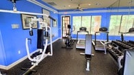 This fitness center in Dallas, Texas, offers a dedicated space for residents or members to maintain an active lifestyle. The room is finished with bold cobalt blue walls accented by white trim, creating a vibrant and energetic environment. Durable rubber flooring lines the entire space, providing a safe, slip-resistant surface ideal for a range of workout activities.

The facility is equipped with a multi-station cable and weight machine, a dedicated workout bench, an elliptical trainer, and multiple treadmills, accommodating both strength training and cardiovascular exercise. A wall-mounted mirror near the strength equipment allows users to monitor their form during workouts.

Natural light filters into the room through windows fitted with horizontal blinds, offering views of surrounding greenery and contributing to an open, inviting feel. Recessed lighting and a ceiling fan ensure the space remains well-lit and properly ventilated during use. An illuminated emergency exit sign reflects attention to safety standards. Overall, the fitness center presents a clean, functional, and well-maintained environment suited for a variety of exercise routines.