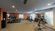 Located at 3000 Greenridge Dr in Houston, Texas, this fitness center offers a comprehensive and well-appointed workout facility on a single floor. The space is designed to accommodate a wide range of exercise needs, featuring a thoughtful layout that maximizes usability and comfort.

The main workout area is outfitted with multiple treadmills, elliptical machines, and a stationary bike, providing ample options for cardiovascular training. A multi-station cable weight machine and a dedicated free weights section — complete with a tiered dumbbell rack and weight bench — cater to strength training. Stability balls and yoga mats are also available for flexibility and functional fitness exercises.

Full-length mirrors line one wall, visually expanding the space and allowing users to monitor their form during workouts. Large windows with horizontal blinds line multiple walls, allowing natural light to filter in and creating an open, airy atmosphere. Recessed lighting and ceiling fans ensure the space remains well-lit and comfortable throughout use.

The interior features warm terracotta-toned accent walls that contrast with neutral gray tones, contributing to an energetic yet welcoming ambiance. A wall-mounted television is positioned for easy viewing during cardio sessions. The durable, patterned tile flooring throughout is both practical and easy to maintain, rounding out a fitness facility that is functional, accessible, and well-suited to an active community.