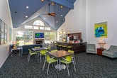 Located at 495 Coachman Dr in Troy, Michigan, this single-floor community space is thoughtfully designed to offer both functional and social areas for its occupants. The layout reveals a generously scaled great room anchored by dramatically high ceilings and large arched windows that flood the interior with natural light, creating an open and airy atmosphere throughout.

The main gathering area features a combination of casual seating arrangements, including upholstered lounge chairs and modular tables with contemporary chartreuse seating, ideal for social interaction or quiet relaxation. A dedicated game area with a pool table is accessible via an open pass-through, adding a recreational dimension to the space. French doors provide connectivity between zones, maintaining an open flow while allowing for defined areas.

A kitchenette with dark cabinetry, granite countertops, and full appliances including a microwave and coffee maker supports everyday convenience. A leasing center is also incorporated within the space, clearly marked and accessible via French doors. Throughout, bold contemporary artwork animates the white walls, while a consistent patterned carpet in dark tones grounds each area cohesively. The overall ambiance is polished, welcoming, and purposefully designed for community living.