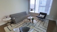 This single-floor space showcases a thoughtfully designed living area with a distinctly modern aesthetic. Large floor-to-ceiling windows flood the room with natural light while framing views of the surrounding urban landscape, giving the space an open and connected feel. The living area centers around a low-profile gray upholstered sofa paired with a sleek black leather accent chair, both positioned atop a textured cream area rug with subtle geometric patterning. A set of round, tiered coffee tables with metallic accents anchors the seating arrangement, providing functional surface space while complementing the room's contemporary design language. An arc floor lamp with a chrome finish adds both task lighting and a sculptural element to the corner of the room. Additional modern seating, including wire-frame accent chairs, contributes to the space's layered and curated character. The neutral color palette—anchored by soft grays, whites, and blacks—creates a cohesive and sophisticated atmosphere throughout. The combination of clean lines, metallic details, and carefully selected furnishings reflects a refined, mid-century modern-inspired interior design sensibility well-suited to urban living.