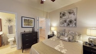 This single-floor residential space offers a thoughtfully designed living environment highlighted by a cozy and elegantly furnished bedroom. The bedroom features soft, neutral-toned walls that create a calm and inviting atmosphere, complemented by plush carpeted flooring underfoot. A ceiling fan with a light fixture is mounted overhead, ensuring year-round comfort and air circulation. The room is generously sized, providing ample space for a large bed, a dark-finished dresser with multiple drawers, and bedside nightstands with warm accent lighting. Decorative wall art featuring botanical motifs adds a refined touch to the space. A standout feature of the bedroom is the walk-in closet, accessible through bifold doors, which offers substantial storage with built-in hanging rods and shelving — a practical and desirable amenity. The overall ambiance of the space is warm, residential, and move-in ready, with a cohesive neutral color palette of beige and gray tones that lend a timeless, understated elegance. The combination of functional storage, comfortable furnishings, and tasteful décor makes this a well-balanced and livable space suited for everyday comfort.