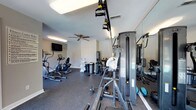 Located at 1 Westchase Drive in Charleston, South Carolina, this fitness center offers a well-equipped, single-floor workout facility designed to support a range of exercise routines. The space features durable speckled rubber flooring throughout, providing both safety and comfort during workouts.

The facility is outfitted with a diverse selection of equipment, including Matrix elliptical trainers, stationary bikes, and a VR1 cable machine for strength training, along with additional weight and resistance apparatuses. A wall-mounted flat-screen television adds convenience for those who enjoy entertainment during their sessions.

Ceiling fans are positioned throughout the room to maintain comfortable airflow, complementing the overhead fluorescent lighting. Full-length mirrors line one wall, enhancing the sense of space and allowing users to monitor their form. Large French-style windows and glass-paneled doors allow ample natural light to filter into the room, creating a bright and inviting atmosphere with views of the surrounding outdoor greenery.

The neutral-toned walls and clean layout contribute to a functional, no-frills environment focused on usability. Towel and accessory stations are conveniently placed within the space. Overall, the fitness center provides a practical and well-maintained setting for residents to pursue their health and wellness goals.