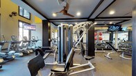 This fitness center is thoughtfully designed to support a comprehensive workout experience. The space features a bold, modern aesthetic with dark trimmed coffered ceilings, recessed lighting, and a ceiling fan that keeps the environment comfortable during intense training sessions. A yellow accent wall adds a vibrant, energetic touch to the overall design.

The strength training area is well-appointed with a multi-station cable machine and a variety of selectorized equipment, allowing users to target multiple muscle groups. A dedicated free weights section lines one wall, complete with medicine balls and a full rack of dumbbells, all reflected in large floor-to-ceiling mirrors that enhance the sense of space and allow for proper form checks.

The cardio zone is visible along the perimeter and includes several treadmills and upright cardio machines, positioned near windows that allow for natural light. A wall-mounted television provides entertainment during cardio sessions. The flooring throughout is a durable, textured gym-grade surface suited for both equipment use and functional fitness activities. Overall, this fitness center offers a well-organized, fully functional environment for a wide range of training needs.