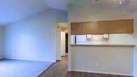 This single-floor residence offers a thoughtfully arranged open layout that maximizes the sense of space throughout. The living area is anchored by striking vaulted ceilings that add volume and an airy feel to the room, with carpet flooring providing a comfortable, warm foundation. Transitioning into the main corridor and dining area, durable hardwood-style flooring takes over, creating a clean visual contrast between the two zones.

The kitchen is positioned along one wall and features a pass-through counter with overhead wood-finish cabinetry, offering both a functional workspace and a convenient connection to the adjacent living and dining areas. White appliances, including a refrigerator and additional kitchen equipment, are neatly integrated into the space. A hallway visible from the main living area leads toward a bedroom with what appears to be a spacious walk-in closet, offering practical storage solutions.

The neutral color palette throughout — soft whites and warm beiges — creates a calm, cohesive ambiance that allows natural light to reflect and brighten the interior. The open sightlines between rooms enhance the overall flow of the floor plan, making the space feel connected and easy to navigate. The combination of vaulted ceilings, open-concept design, and functional kitchen layout makes this a practical and welcoming single-floor home.