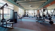 This fitness center is a comprehensively outfitted facility designed to support a full range of workout routines. The space features an open, airy layout beneath an exposed beam ceiling with a teal finish, complemented by large windows along one wall that flood the floor with natural light and offer views of the surrounding exterior. Ceiling fans provide comfortable air circulation throughout.

The cardio zone is well-stocked with treadmills, stair climbers, and elliptical machines arranged to take advantage of the natural light from the windows. A dedicated strength training area houses a multi-station cable machine, a lat pulldown/cable system, and an adjustable bench, while a separate free weight section offers a full rack of dumbbells alongside additional benches and a wall-length mirror that enhances the sense of space.

A functional training area is set apart with a multi-functional rig, medicine balls, a plyo box, and resistance accessories for dynamic and cross-training workouts. Bold, large-format motivational murals line the walls with energizing phrases, adding an inspiring atmosphere to the environment. Wall-mounted televisions are positioned throughout for entertainment during workouts. The flooring consists of durable rubber tile, providing safety and support across all exercise zones.