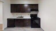 This single-floor unit offers a practical, well-organized layout with distinct living, sleeping, kitchen, and bathroom areas. The kitchen is equipped with rich, dark espresso-toned cabinetry spanning both upper and lower sections, providing ample storage. Light-toned countertops contrast the dark cabinetry, and the space includes a built-in sink, black electric range with overhead ventilation, and a dishwasher. Light wood-style flooring runs throughout the kitchen area, contributing to a clean, functional aesthetic.

The living area is generously proportioned, featuring neutral carpet flooring and cream-toned walls that create a bright, open atmosphere. A structural beam detail adds architectural character, and a thermostat is conveniently positioned on the wall. Two side-by-side windows allow natural light to filter in, offering views of the surrounding exterior. The bedroom areas are similarly finished with plush carpet and neutral walls, providing a calm, comfortable environment with closet access.

The bathroom features white subway tile wainscoting, a dark-framed mirror, a modern brushed nickel faucet set into a white vanity with dark cabinetry below, and wood-style flooring, delivering a clean and updated appearance throughout.