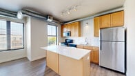 Located at 2401 Chicago Ave in Minneapolis, MN, this one-bedroom, one-bathroom unit offers a thoughtfully designed open-concept layout across a single floor. The combined kitchen and living area forms the heart of the home, characterized by loft-inspired exposed metal ductwork running along the ceiling and track lighting that adds an industrial yet modern aesthetic. Large windows allow generous natural light to fill the space, with views of the surrounding Minneapolis neighborhood.

The kitchen is well-appointed with warm-toned wood cabinetry, a central island featuring a crisp white countertop, stainless steel appliances including a gas range, built-in microwave, and refrigerator, plus a stainless steel sink with a modern faucet. The living area extends seamlessly from the kitchen, featuring durable wood-style flooring throughout.

The bedroom is a comfortable, carpeted retreat accessed through a sleek glass-paneled door, offering a quiet separation from the main living space. The bathroom features a generous vanity with white countertops, warm wood cabinetry, a well-lit mirror, and a bathtub/shower combination with wood-style flooring. Additional highlights include a den, in-unit laundry, a hallway, and multiple closets providing ample storage throughout.