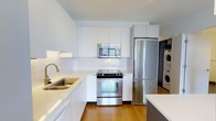 This single-floor residence offers a thoughtfully designed layout within a compact yet efficient footprint. The kitchen serves as a standout feature, showcasing a sleek, modern aesthetic with flat-front white cabinetry that extends to the ceiling, maximizing storage capacity. Stainless steel appliances — including a full-size refrigerator, range with oven, and over-the-range microwave — provide a polished, cohesive look. White subway tile backsplash adds a clean, classic touch, while light wood flooring grounds the space with warmth. A double-basin stainless steel sink sits along the counter, complementing the white quartz or composite countertops that wrap the kitchen perimeter. Notably, a stacked washer and dryer unit is conveniently located just off the kitchen, offering in-unit laundry — a practical and desirable feature. The overall design palette of white, stainless steel, and natural wood tones creates a bright, contemporary ambiance throughout. The residence is well-suited for those seeking a modern, low-maintenance living space with efficient use of every square foot.